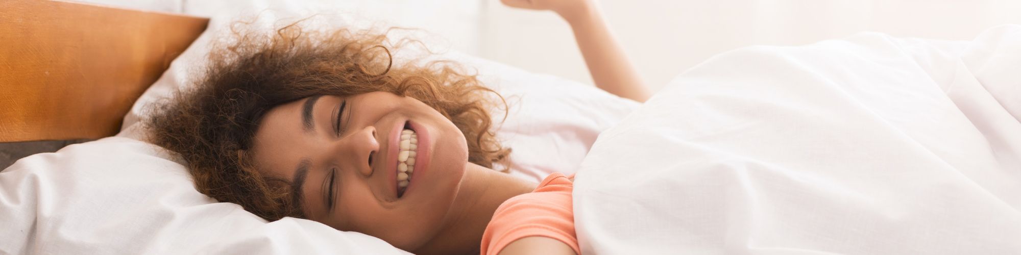 A person is lying in bed, smiling with eyes closed, stretching their arm upwards under a white blanket, and appearing happy and relaxed.