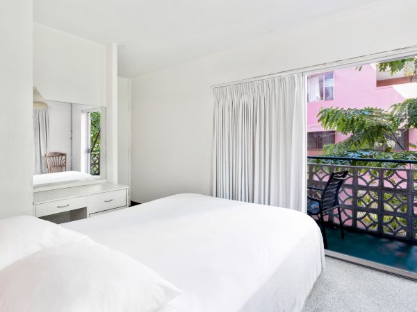 A bright bedroom with a double bed, a dressing table, and large window with curtains leading to a balcony overlooking greenery.