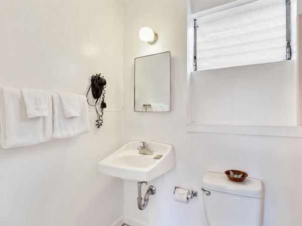 A small bathroom with a sink, mirror, towels, a light fixture, and a toilet beneath a window with a blind.