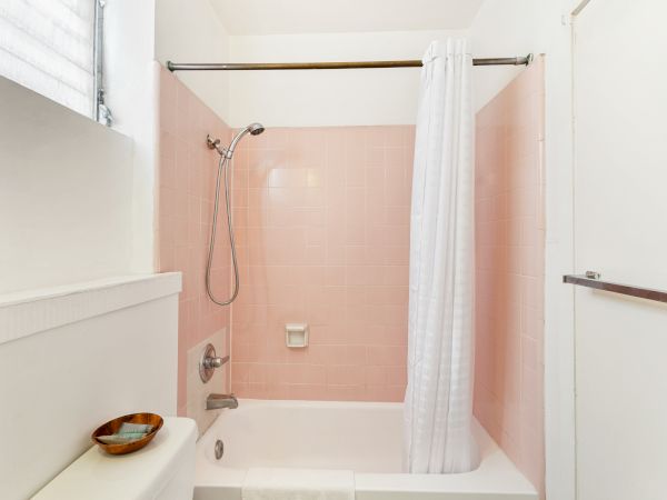 A bathroom with a pink tiled bathtub and shower, white curtain, window, and a small bowl on a ledge.