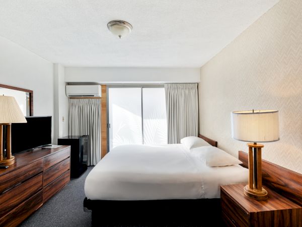 The image shows a hotel room with a bed, lamps, a dresser, and a TV, featuring neutral colors and a window with curtains.