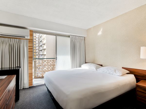 A hotel room with a large bed, nightstand, lamp, TV, and air conditioner, featuring a balcony view with curtains partly open.