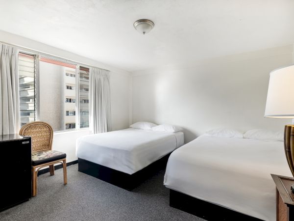 The image shows a hotel room with two beds, a chair by a window, and a lamp on a side table. The atmosphere is bright and simple.