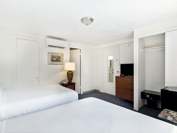 The image shows a hotel room with two beds, a TV, a lamp on a nightstand, and an open closet with hangers and a safe.