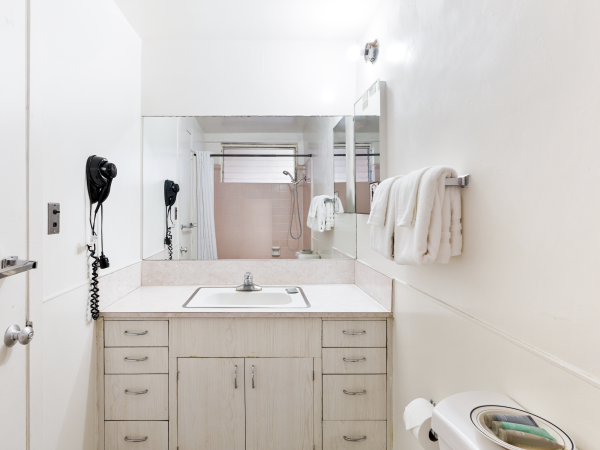 The image shows a clean bathroom with a sink, mirror, hairdryer, towels, and a toilet, all in a white and minimalist design.