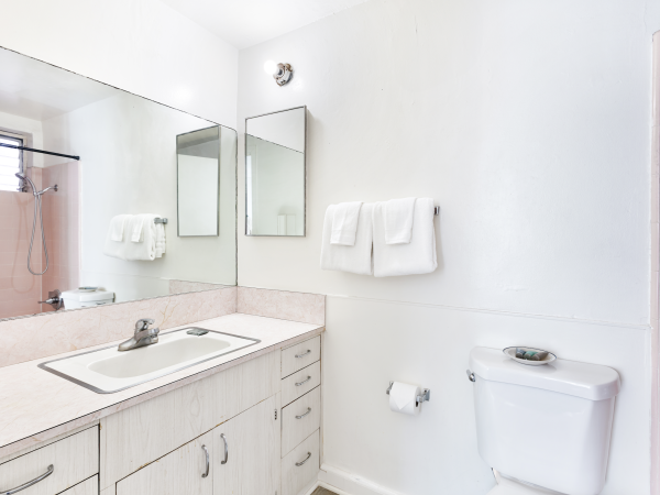A clean, bright bathroom with a large mirror, sink, white towels, and a toilet. The design is simple and minimalist.