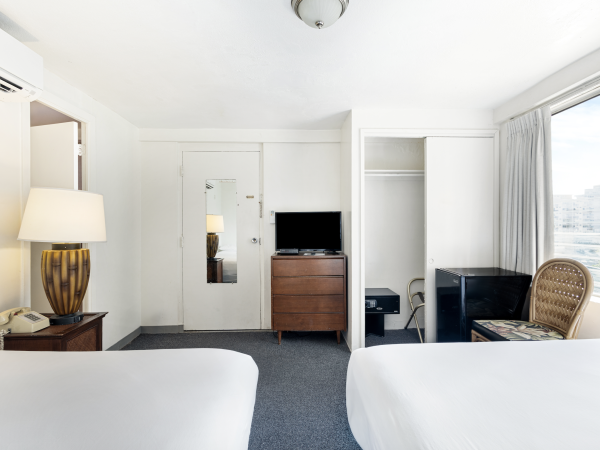 The image shows a hotel room with two beds, a lamp, TV, a chair, closets, and a window, all in a bright setting.