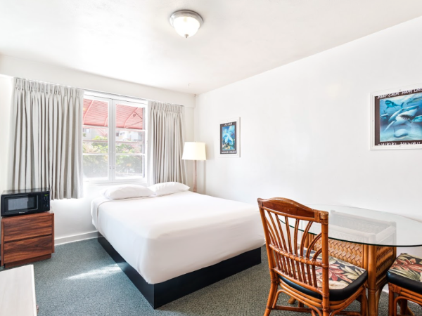 A tidy hotel room with a bed, table and chairs, a window, a lamp, and wall art. A microwave is on a small table near the bed.