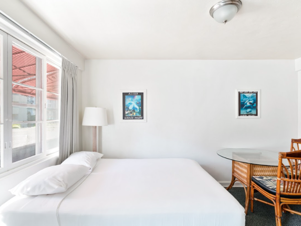 A bright room with a bed, two framed artworks, a lamp, and a table with chairs by the window.