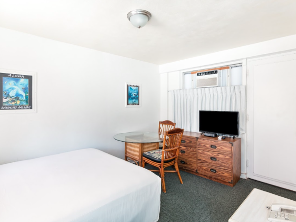 A small room with a bed, wooden dresser, TV, table, chairs, wall art, and an air conditioner beneath a window.