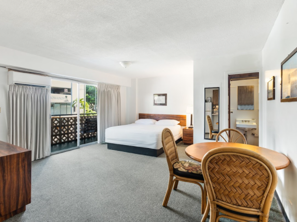 A hotel room with a white bed, table, chairs, TV console, and balcony access, featuring a bright and clean interior.