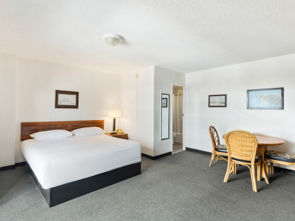 The image shows a hotel room with a bed, nightstand, lamp, and a table with three chairs in a carpeted space.