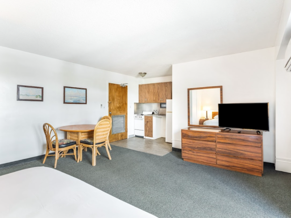 The image shows a room with a bed, table, chairs, TV on a wooden dresser, and a small kitchenette. Walls are decorated with paintings.