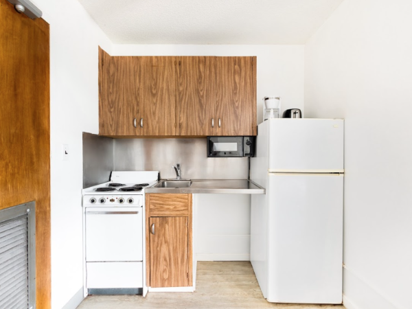 A compact kitchen with a stove, sink, wooden cabinets, countertop, microwave, and refrigerator in a minimalist design.