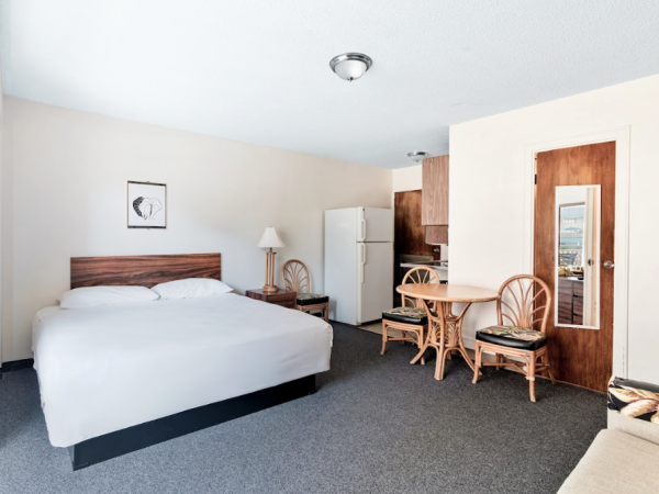The image shows a hotel room with a bed, fridge, small table with chairs, and a wall-mounted lamp on a carpeted floor.