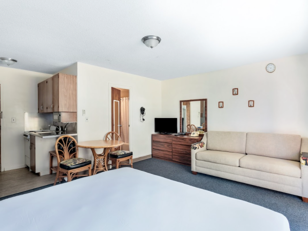 The image shows a hotel room with a bed, sofa, TV on a dresser, a small table with chairs, and a kitchenette.