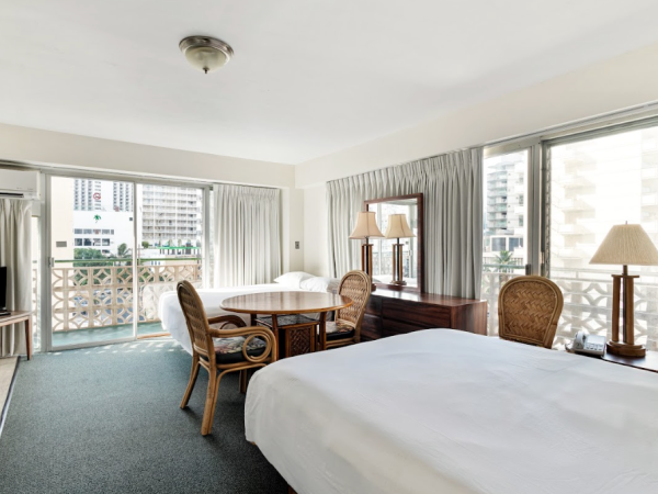 The image shows a hotel room with a bed, a round table with chairs, a TV, and a balcony with sliding glass doors and curtains.
