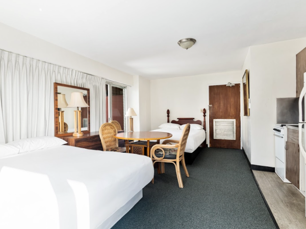 The image shows a hotel room with two beds, a table, chairs, a TV, a fridge, and curtains. The room is well-lit and simply decorated.