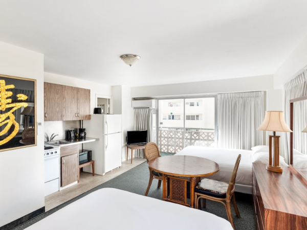 The image shows a hotel room with a kitchenette, two beds, a dining table, chairs, a TV, and a framed painting on the wall.