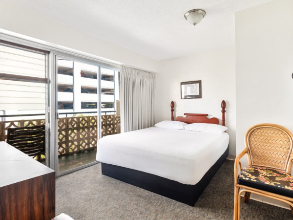 A hotel room with a double bed, chair, dresser, and a balcony view. The room is bright and simply decorated.