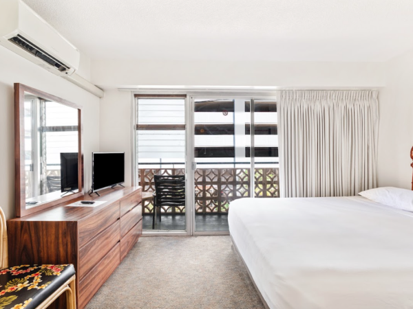 The image shows a neatly arranged hotel room with a bed, TV, dresser, chair, and sliding doors leading to a balcony.