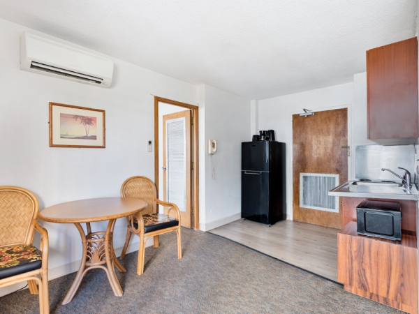 The image shows a simple apartment with a kitchenette, table, chairs, and air conditioning, offering a cozy, minimalistic design.