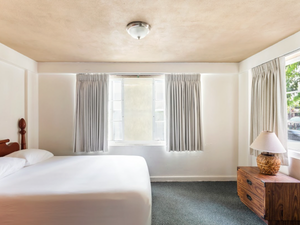 A neatly arranged bedroom with a bed, wooden nightstand with a lamp, window with curtains, and a simple ceiling light fixture.