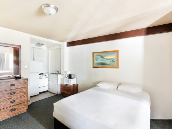 The image shows a small, neatly arranged room with a bed, dresser, kitchen area, and wall art.