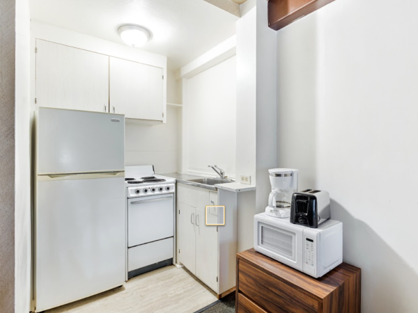 A compact kitchen with a refrigerator, sink, stove, microwave, and coffee maker. White cabinets and wooden shelves complete the setup.