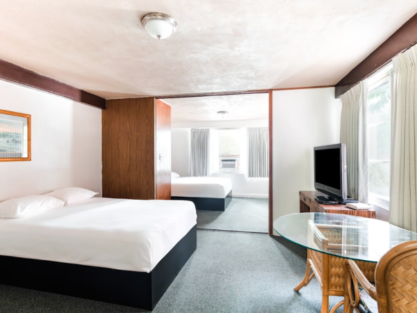 The image shows a hotel room with two beds, a TV, a glass table with chairs, and a large mirrored wall.