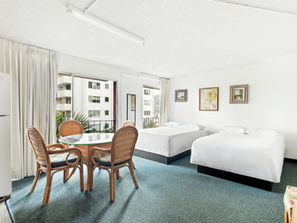 The image shows a hotel room with two beds, a small table with chairs, a refrigerator, and framed artwork on the walls.