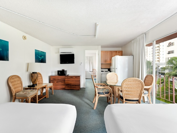 The image shows a hotel room with two beds, wicker furniture, a TV, artworks, and a balcony overlooking greenery and nearby buildings.