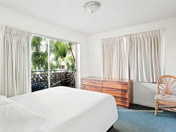 A bright bedroom with a double bed, dresser, chair, and windows showing greenery outside. White curtains and walls complete the look.