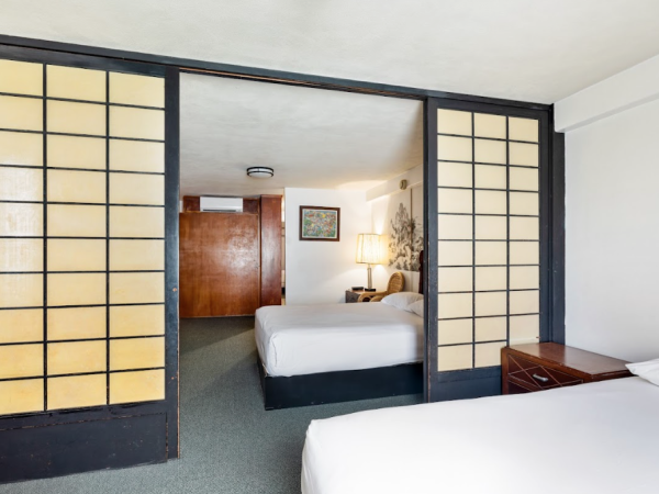 The image shows a room with two beds, a shoji-style sliding door, a bedside table, lamp, and a picture on the wall in a modern interior.