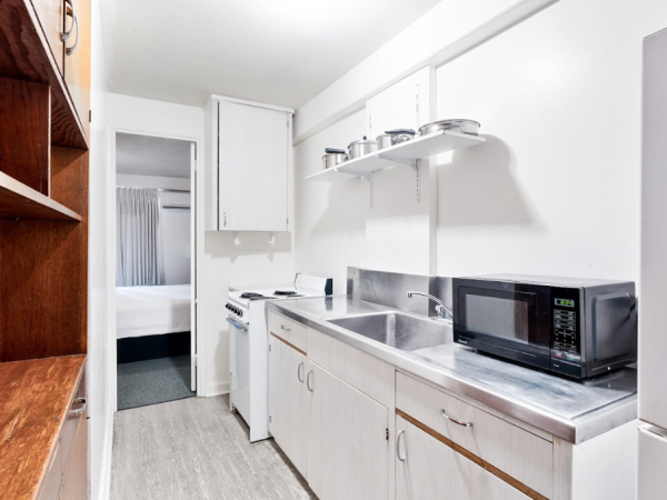 A small kitchen with white cabinets, a microwave, sink, stove, and wooden shelves leads to a room with a bed in the background.