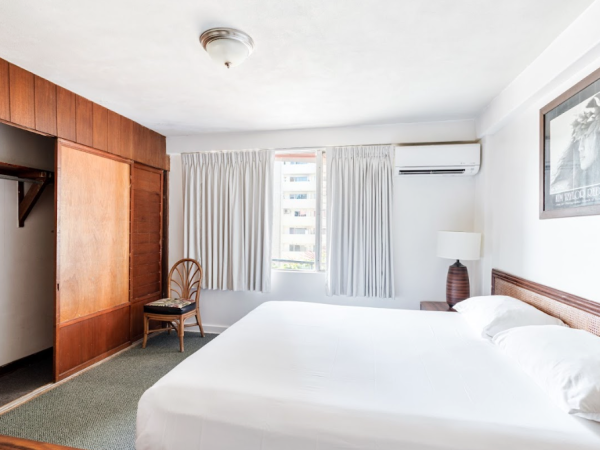 A simple bedroom with a bed, open closet, chair, lamp, and picture. It has white walls, curtains, and a window for natural light.