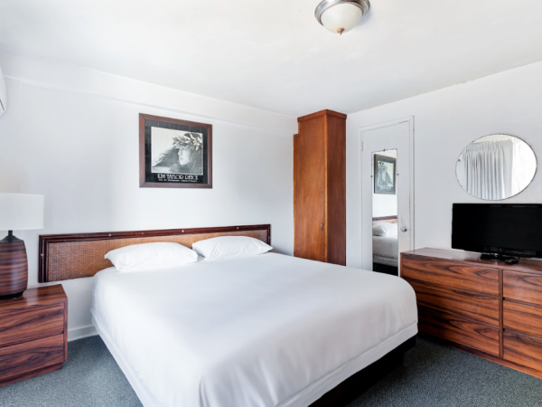 A modern bedroom with a white bed, wooden nightstand, mirror, TV, art above the bed, and an air conditioner on the wall.
