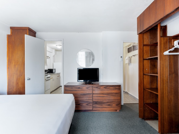 A minimalist hotel room with a bed, wooden furniture, a TV, and an open closet; a doorway leads to a kitchen and bathroom area.