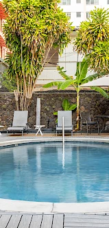 This image shows a round swimming pool surrounded by lounge chairs and umbrellas, with a building and palm trees in the background.