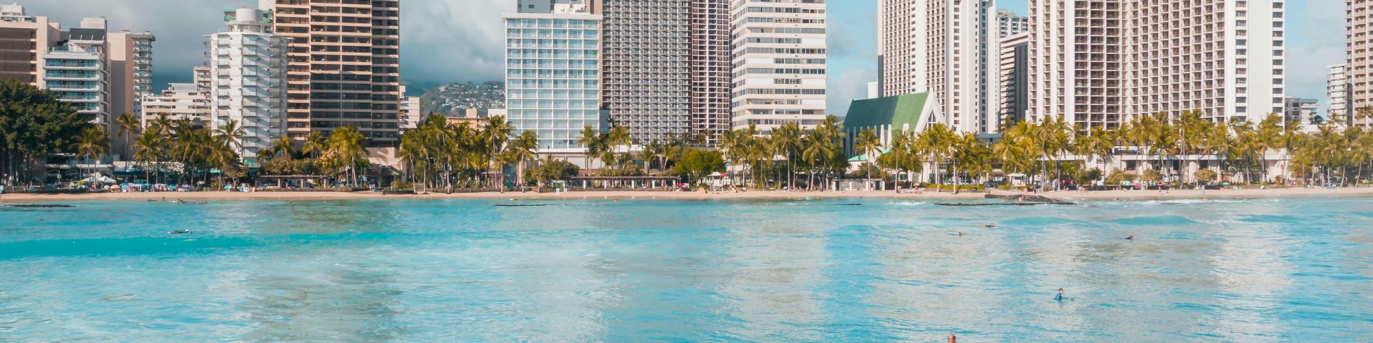 People swimming and riding waves in a bright turquoise ocean with a city skyline of tall buildings along the shore behind them.