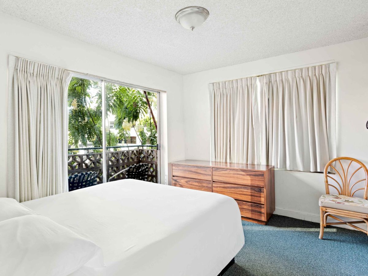 A bright bedroom with a large bed, dresser, chair, and curtains. There's a window overlooking trees and a small balcony outside.
