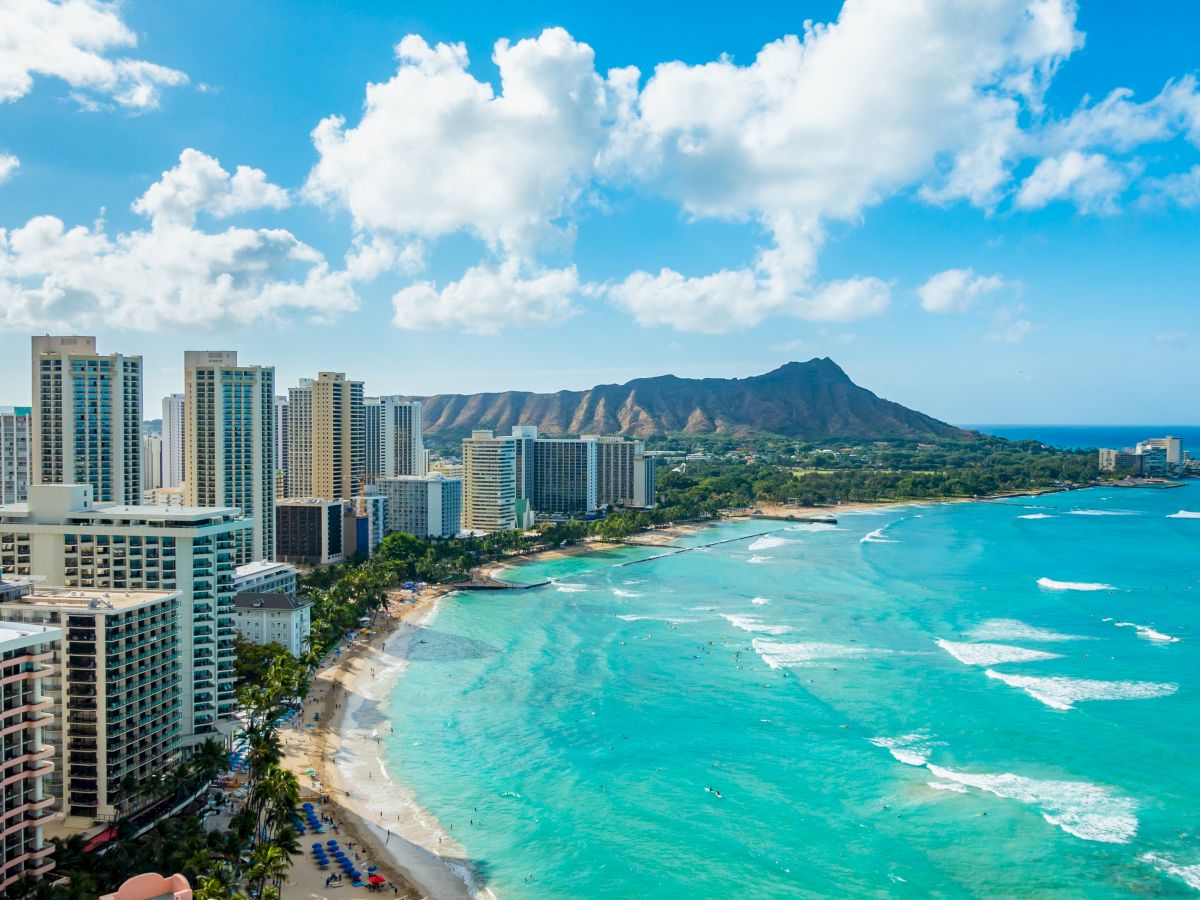 High-rise beachfront cityscape with a turquoise bay, white sandy beach, and a distant mountain backdrop under a bright, partly cloudy sky.