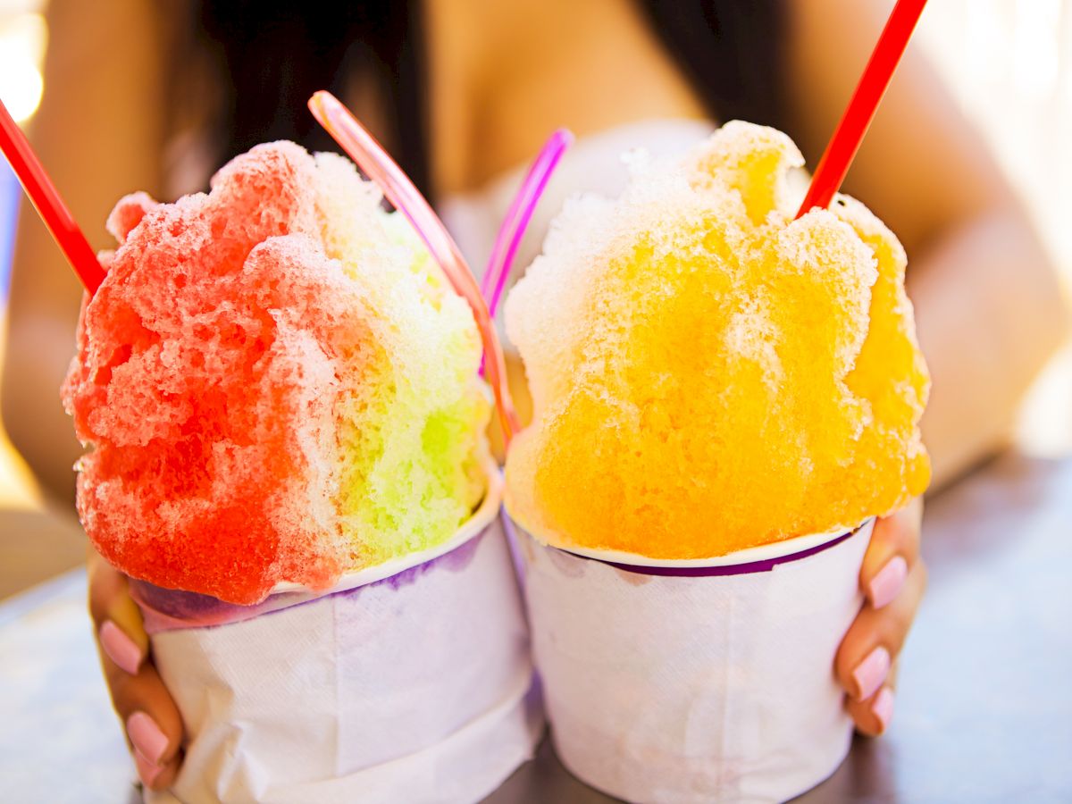 Two colorful shaved-ice desserts in cups with red straws, held by a person, colorful scoops of red/green and yellow/orange.