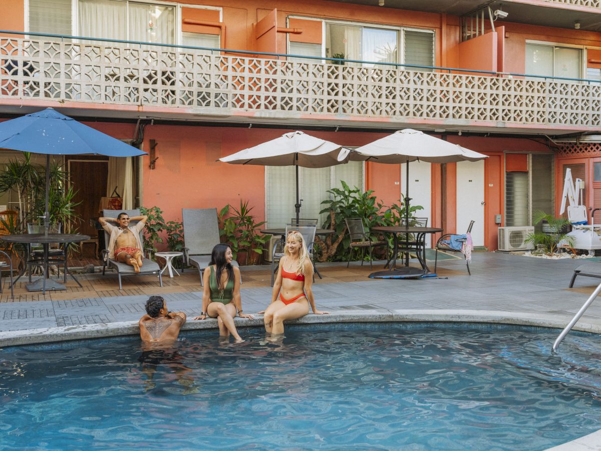 People lounging by a hotel pool under umbrellas, with a peach-colored building featuring balconies in the background.