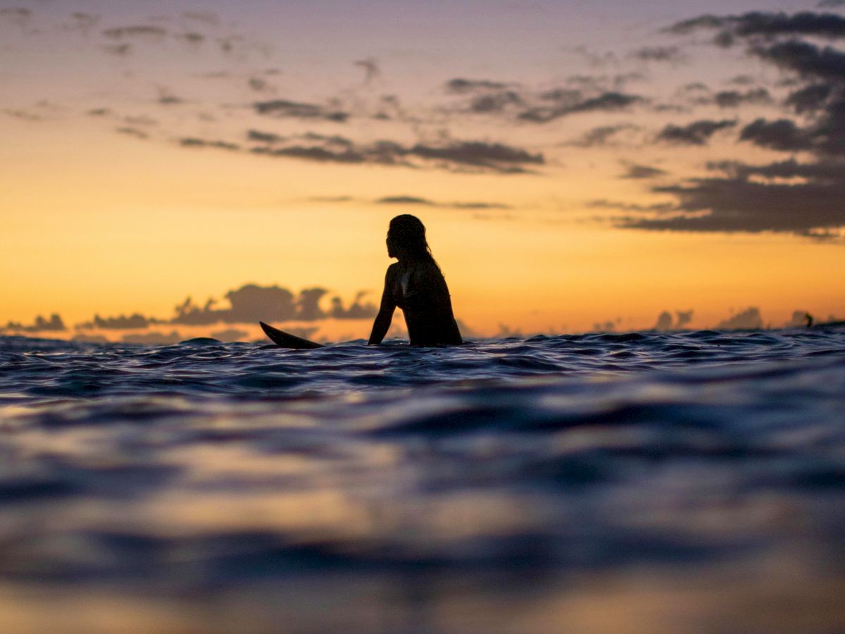 A lone surfer sits on a submerged board at sunset, silhouetted against a orange sky with gentle waves.