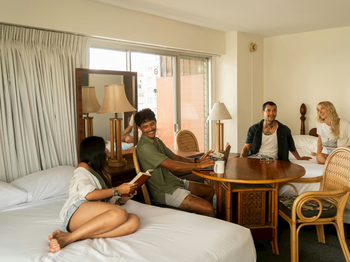 A group of four friends sits around a wooden table in a hotel room, with one person lounging on a bed and a casual, relaxed vibe.