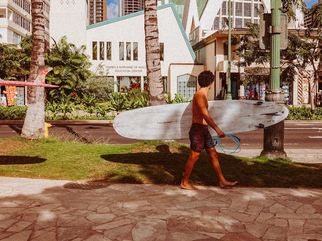 A shirtless man with a surfboard walks along a sunny sidewalk near palm trees and modern buildings.