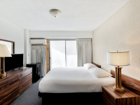 A bright hotel room with a large bed, wooden nightstands, a dresser with a TV, and a sliding glass door leading to a balcony, airy and neat.