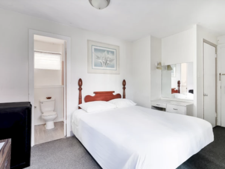 A clean, minimal hotel room with a double bed, white linens, a small vanity, a mirrored door, and an attached bathroom visible in the corner.