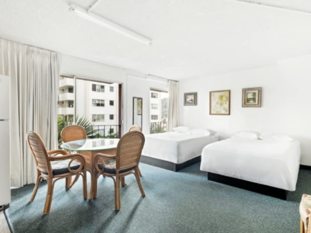 A bright hotel room with two large white beds, a round glass table, four wicker chairs, and light curtains; framed art on the walls.